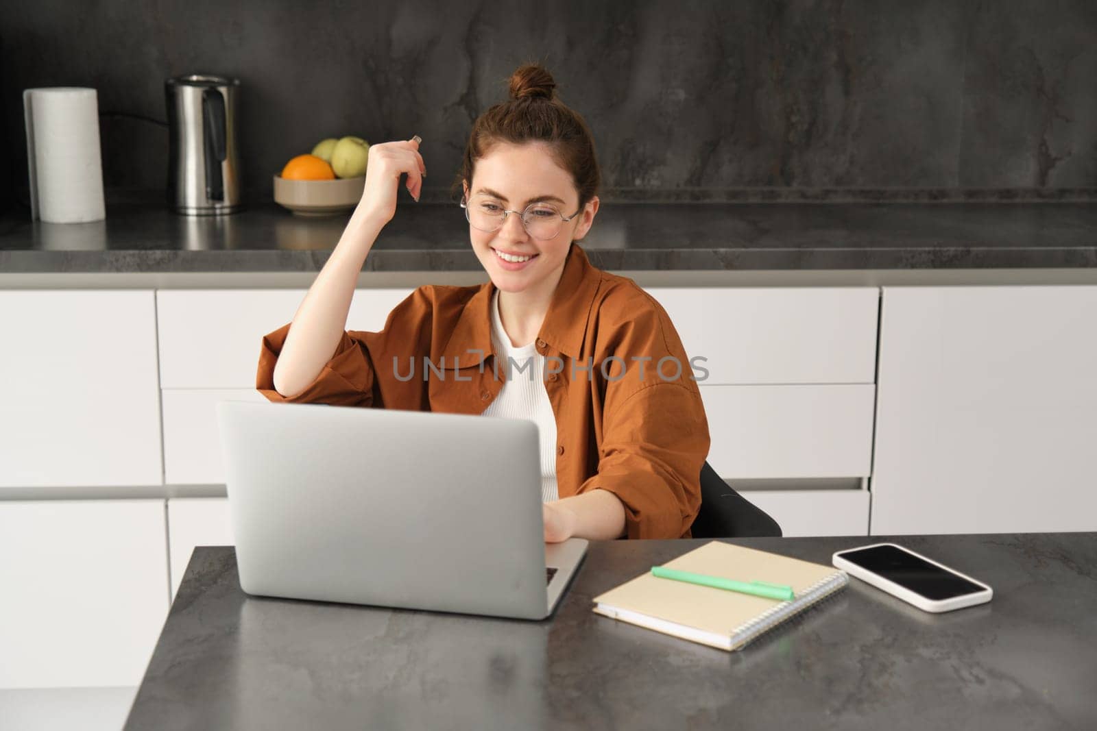 Portrait of young woman, business owner in glasses, sitting with laptop in kitchen, working from home. Student studying, doing homework on computer.