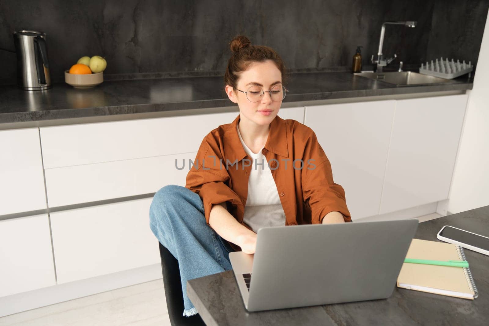 Portrait of young woman, student in kitchen, sitting with laptop, working from home, studying or doing homework, connects to online chat or course from computer.