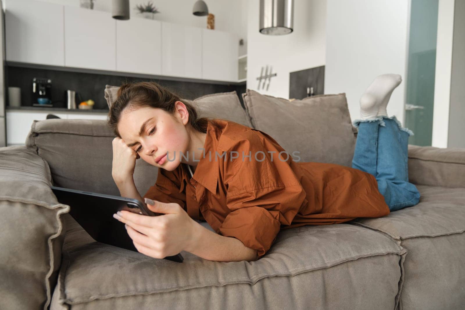 Portrait of woman with sad face, lying on couch at home, resting in living room with digital tablet, frowning and looking frustrated.