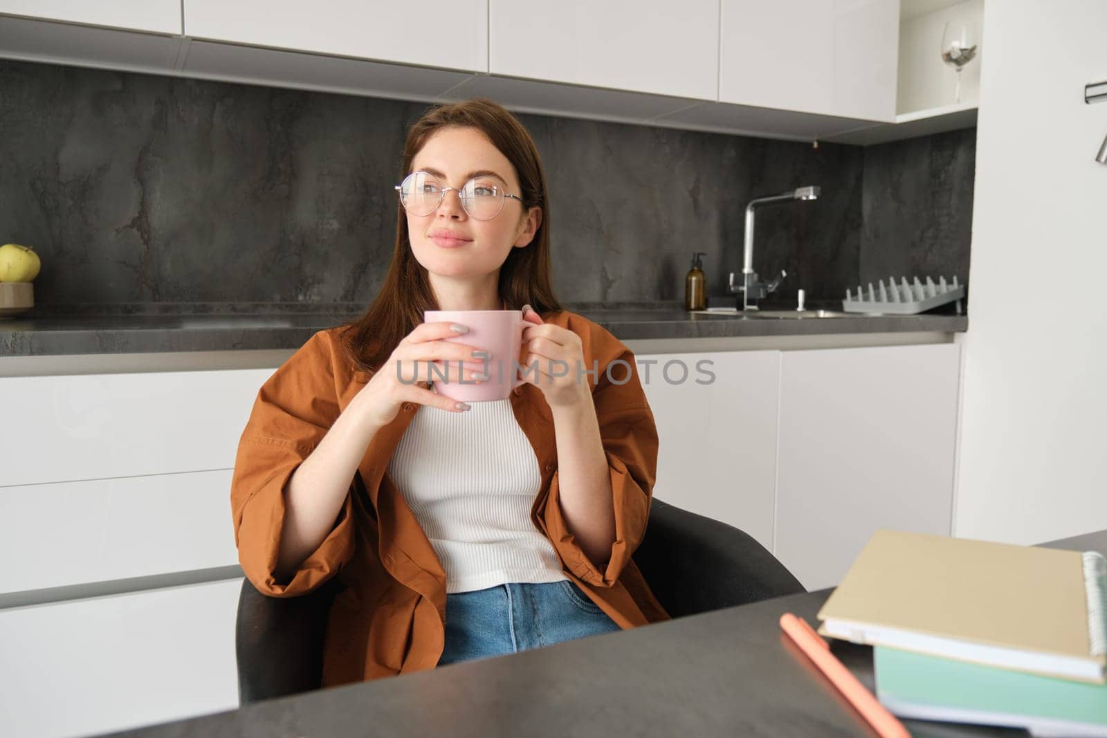 Portrait of young professional, girl freelancer sitting in kitchen and enjoying cup of coffee, sitting with documents, taking break from studying and smiling, looking outside window.