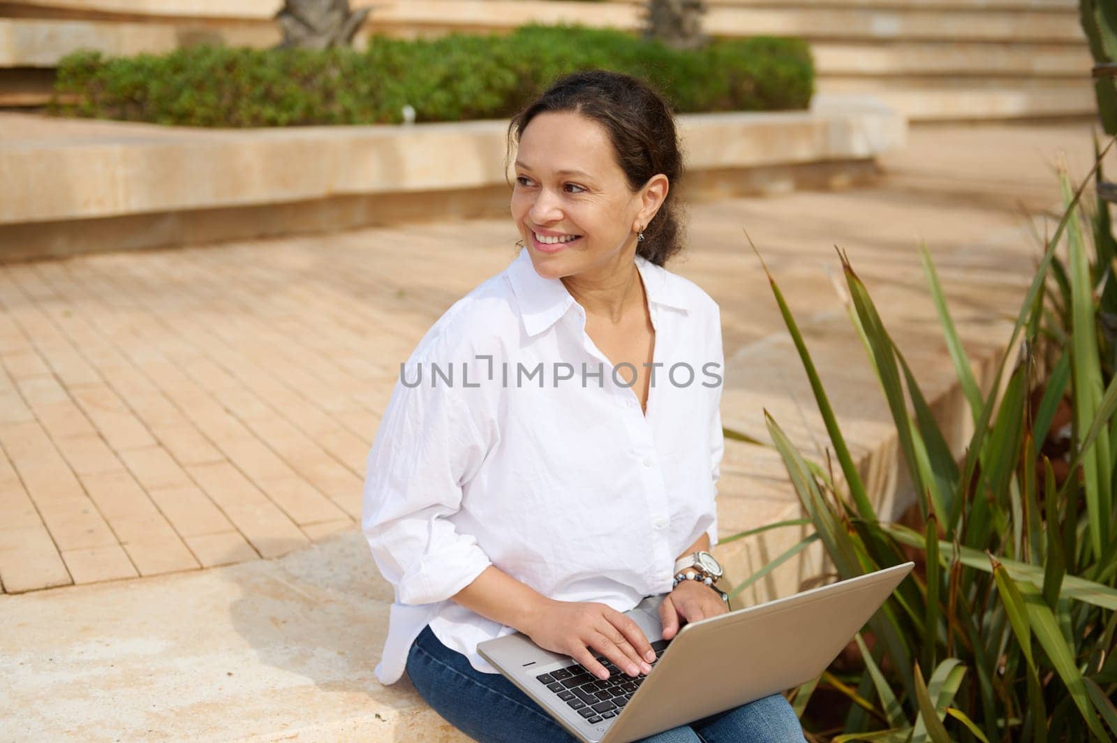Confident female entrepreneur, young adult business woman in casual attire, remotely working on laptop computer, smiling looking aside, planning business startup