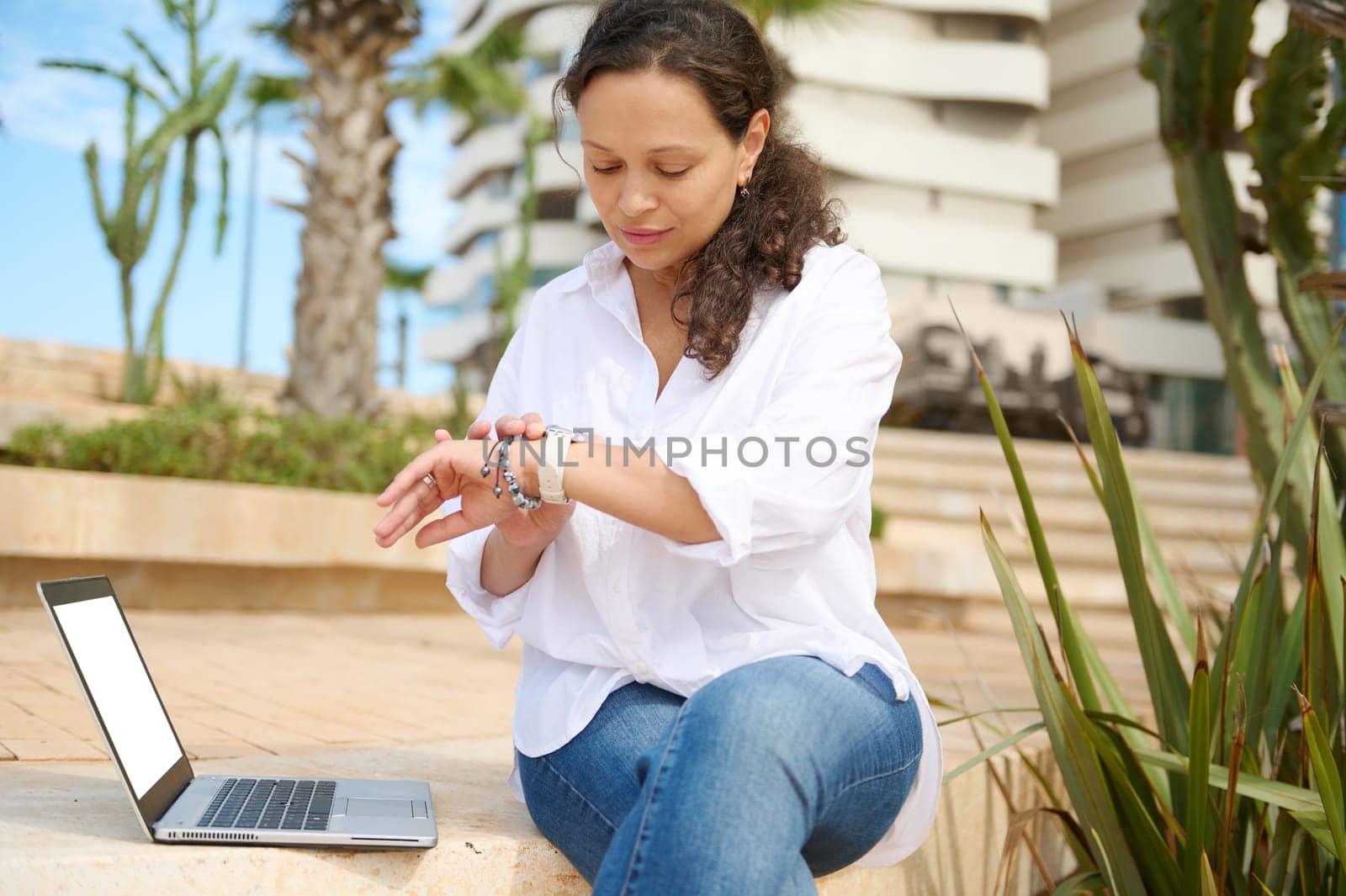 Authentic portrait of a confident multi ethnic young business woman checking time on her wrist watch, online working on laptop outdoor. People. Internet. Innovation. Online communication. Career