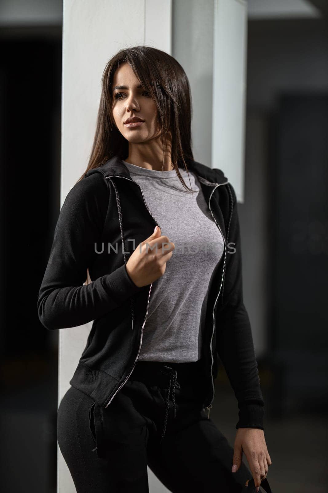 Beautiful fitness model brunette woman in a modern black tracksuit posing in the locker room, demonstrating healthy sexy body and sports fashion