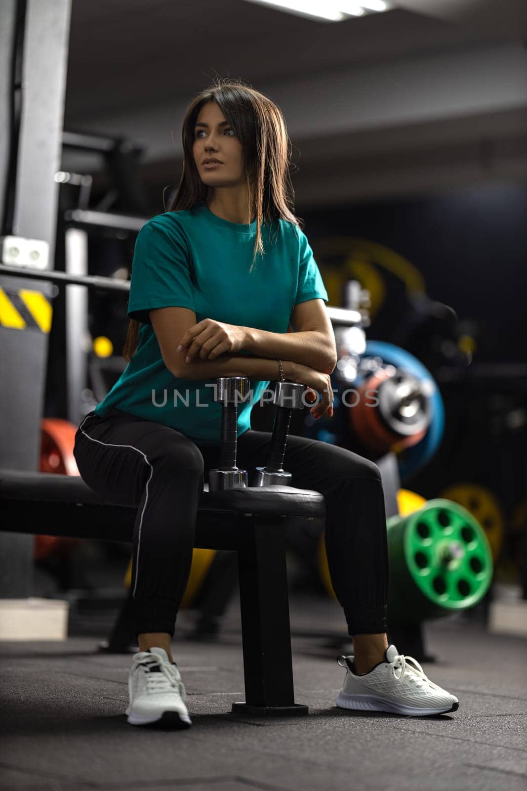Attractive brunette athletic girl in green T-shirt and black sweatpants leans against bench press bar in the gym. Healthy lifestyle, sexy female body and sports fashion