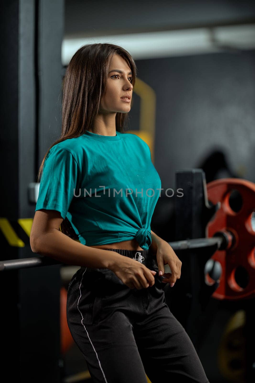 Attractive brunette athletic girl in green T-shirt and black sweatpants leans against bench press bar in the gym. Healthy lifestyle, sexy female body and sports fashion