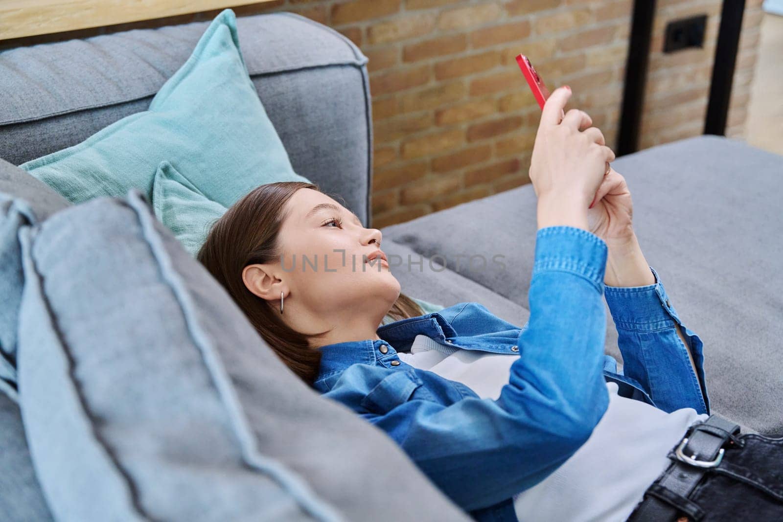 Young smiling happy woman using smartphone, lying on sofa at home in living room. Internet online services, communication, mobile applications, technologies for leisure study work lifestyle