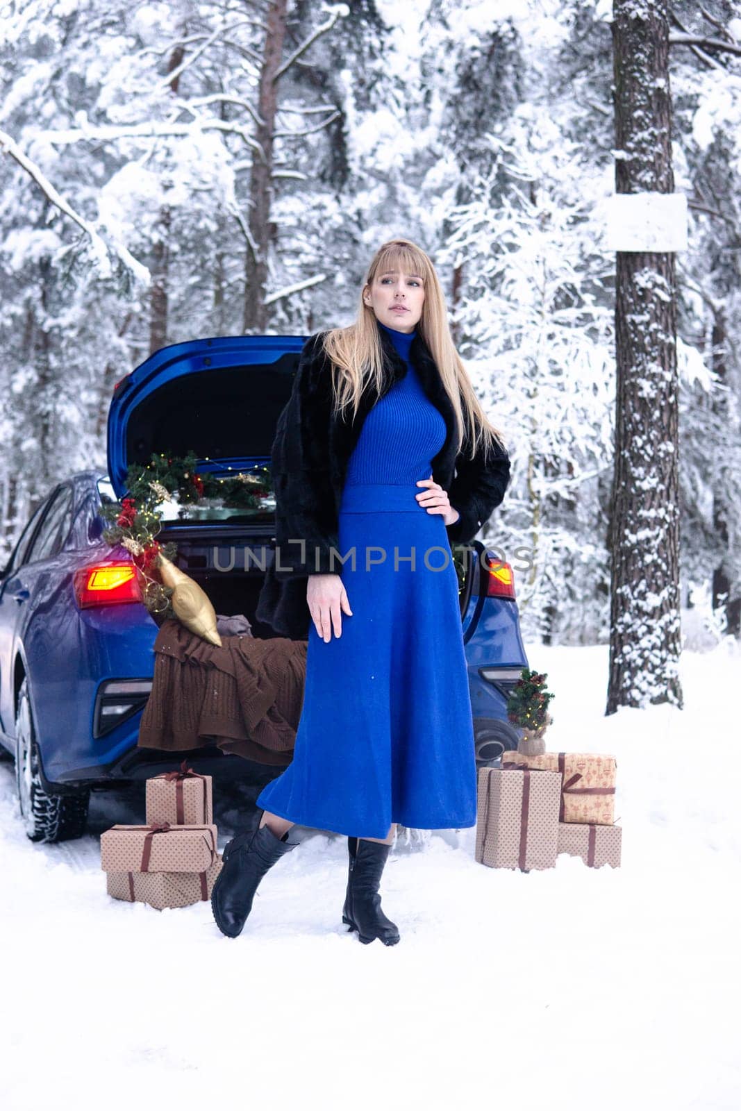 Woman in winter snowy forest in blue dress next to blue car decorated with Christmas decor. Christmas and winter holidays concept