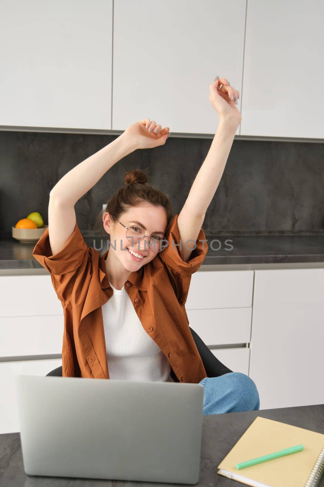 Vertical shot of smiling, lively young woman in glasses, working on remote, sitting at home with laptop, using computer to study online, stretching hands with pleased emotion.