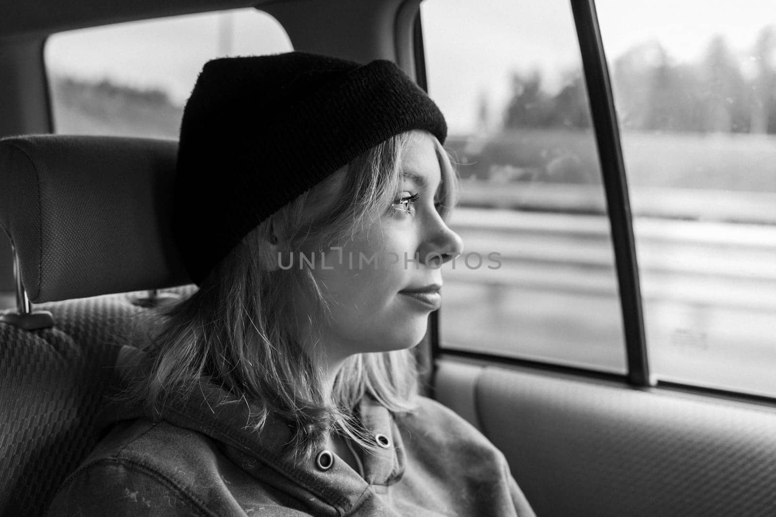 black and white photo. A pretty girl with pink hair in a black knitted hat in the back seat of a car, looking out the window, she is immersed in thoughts about her future, about opportunities. Traveling by car