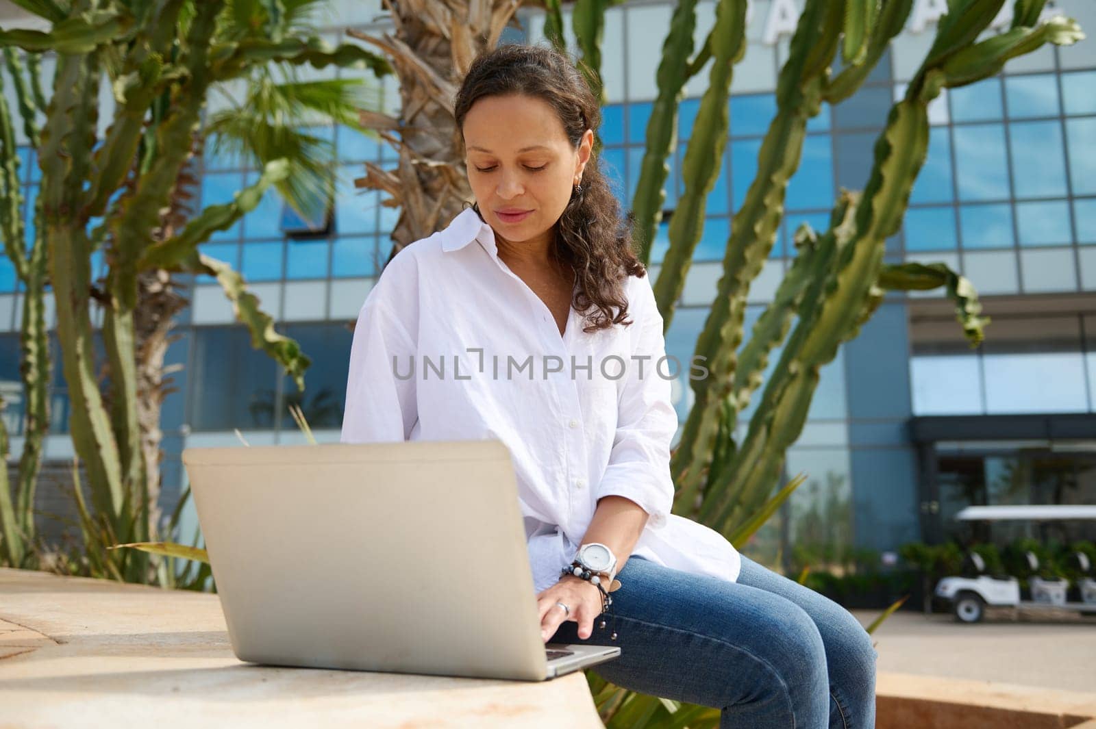 Latin American young woman student in blue jeans and white shirt, typing text on laptop, online working on diploma project sitting outdoor on a stairs, against modern high rise buildings background
