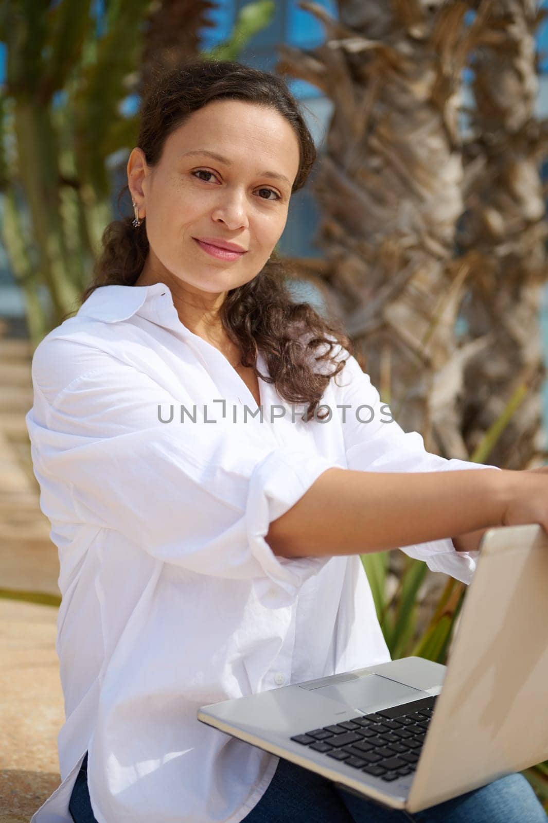 Portrait of a multi ethnic young adult pretty woman, sales manager, real estate agent, business lady, freelance entrepreneur in white casual shirt holding laptop and confidently looking at the camera