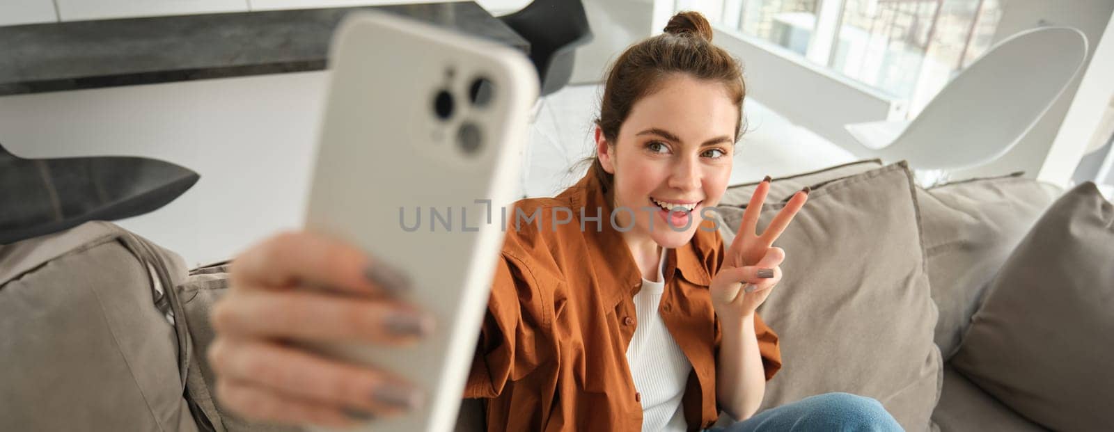 Portrait of beautiful happy girl takes selfies on sofa. Woman posing for photos on her couch, taking pictures for social media, extends hand with smartphone by Benzoix