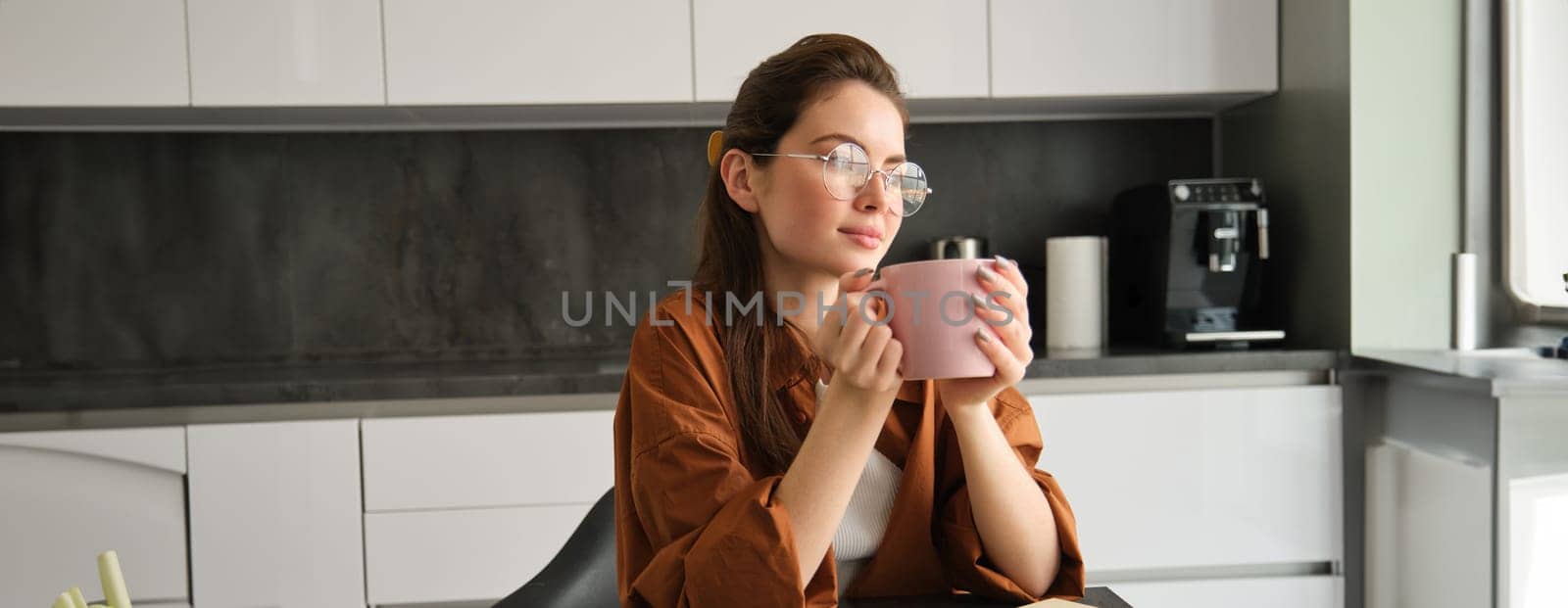 Portrait of young self-employed woman, working from home, taking break for coffee, sitting with documents in kitchen, looking away by Benzoix