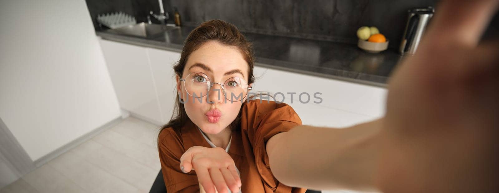Selfie of happy young and carefree woman, taking photo on mobile phone with extended hand, posing and smiling, sitting in kitchen in glasses.