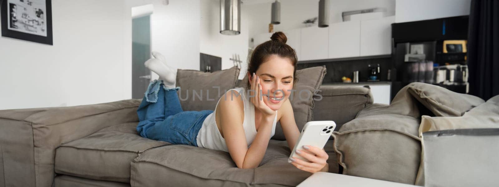 Portrait of smiling, happy young woman resting at home, resting on couch with smartphone, using mobile phone application by Benzoix