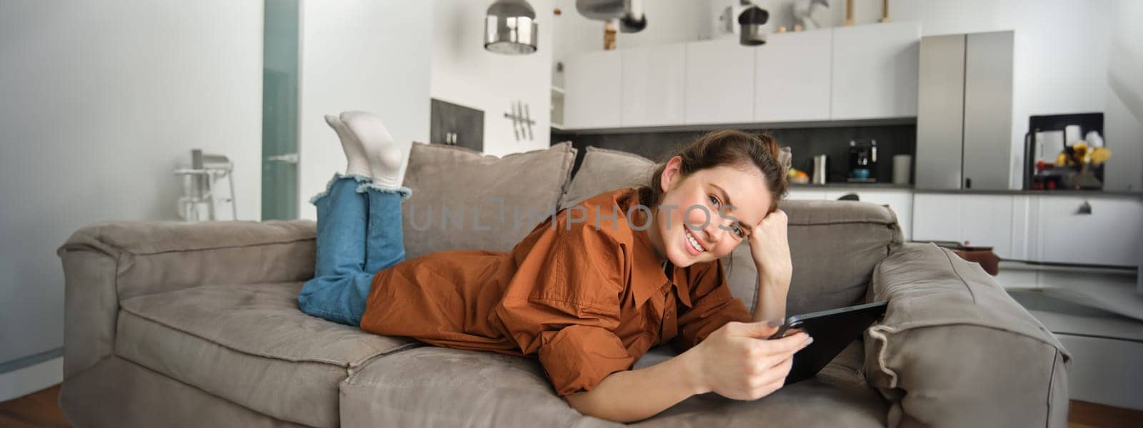 Portrait of smiling, beautiful brunette woman spending time at home, using digital tablet, lying on sofa and looking happy.