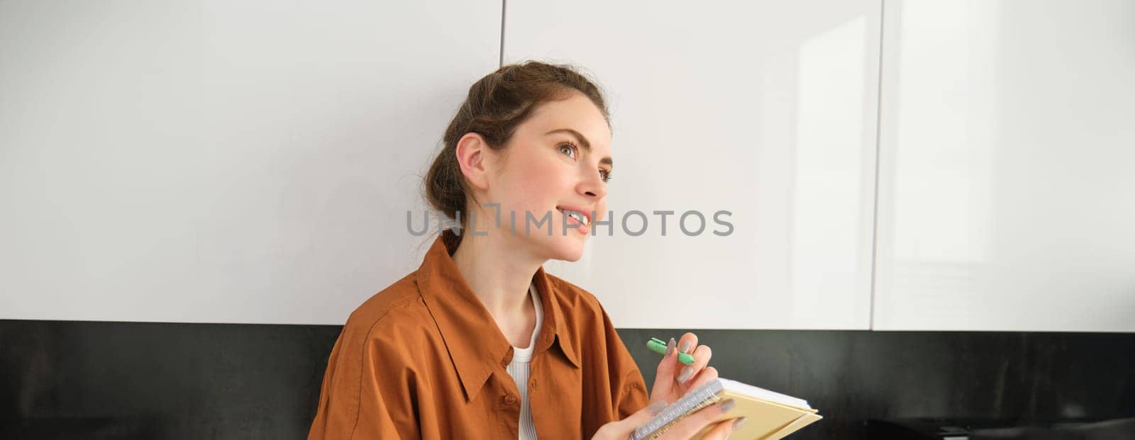 Portrait of woman writing down her ideas in a notebook, holding pen and looking thoughtful, inspired to write in diary, plans her schedule or makes a list for grocery shopping.