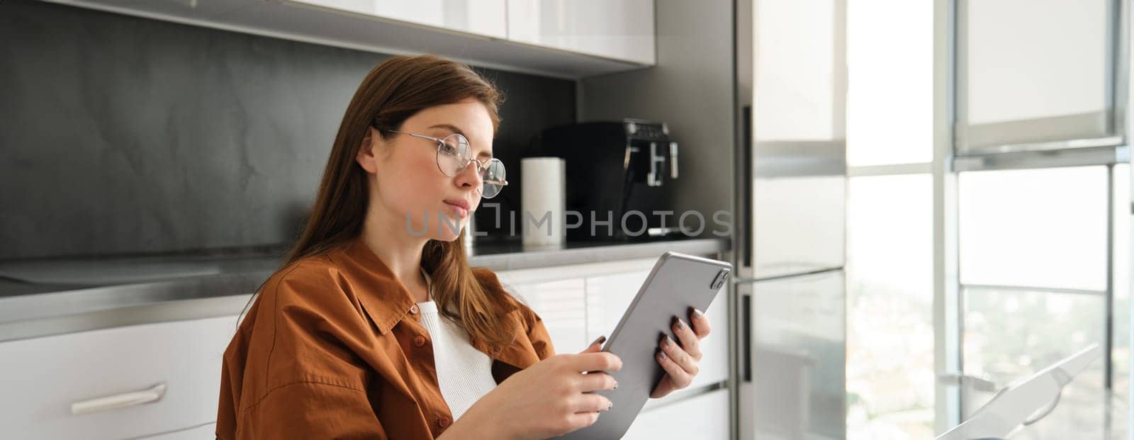 Home and leisure concept. Portrait of beautiful young woman reading on digital tablet, sitting in kitchen, working on remote.