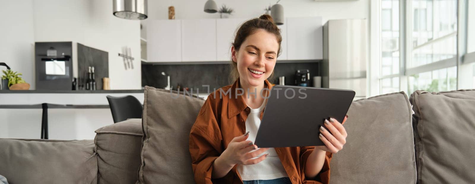 Portrait of young woman sitting on couch in living room, watching movies on digital tablet, reading on device, enjoying weekend at home by Benzoix