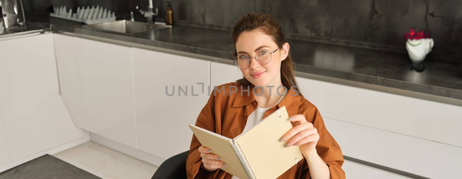Portrait of beautiful young woman studying, student revising at home, doing homework, holding planner, reading her notes and smiling, writing in diary by Benzoix