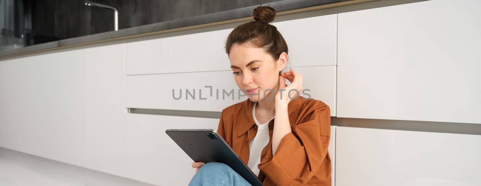 Portrait of pretty young woman at home, sitting on kitchen floor, looking at digital tablet, using gadget, reading on device.