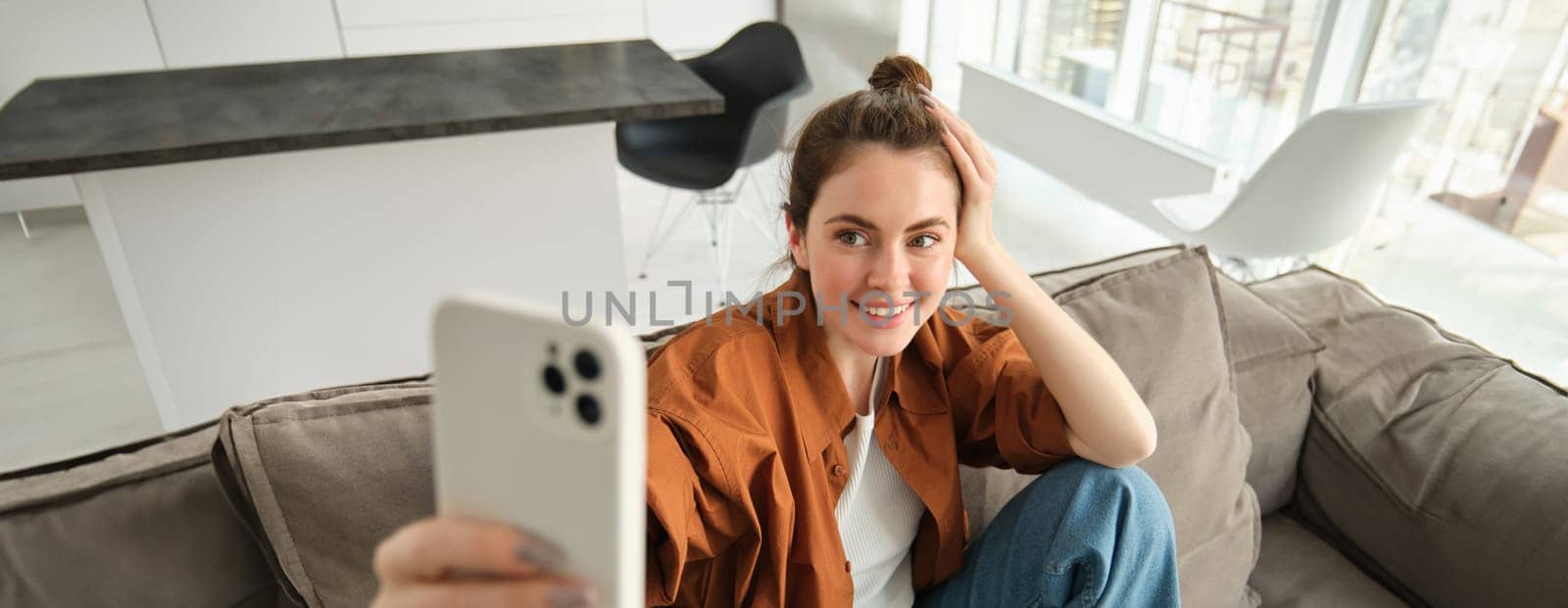 Portrait of stylish, modern young woman taking selfie on smartphone, sitting on couch with mobile phone, taking pictures for her lifestyle blog.