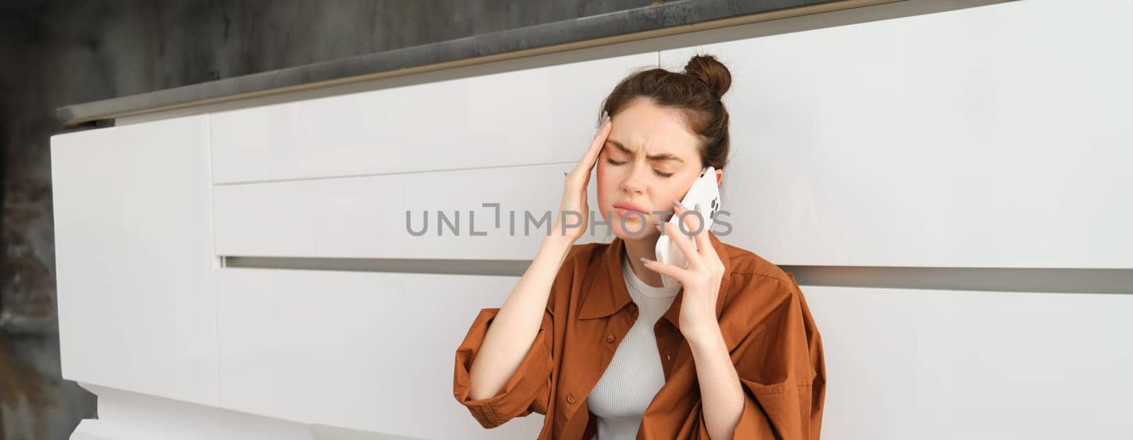 Woman booking appointment with GP over the phone, has headache or migraine, calling in sick, sits on floor at home by Benzoix