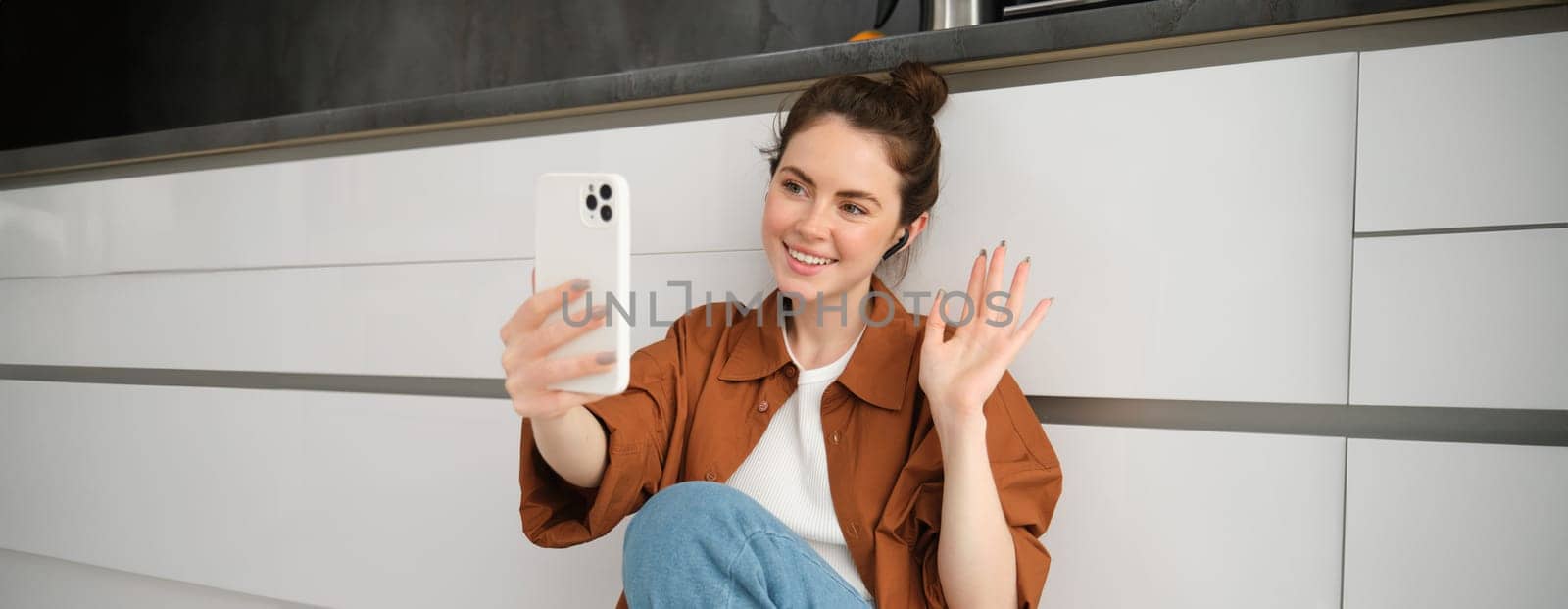 Portrait of happy smiling woman video chats, sits on kitchen floor at home, talking to friend on social media app using smartphone.