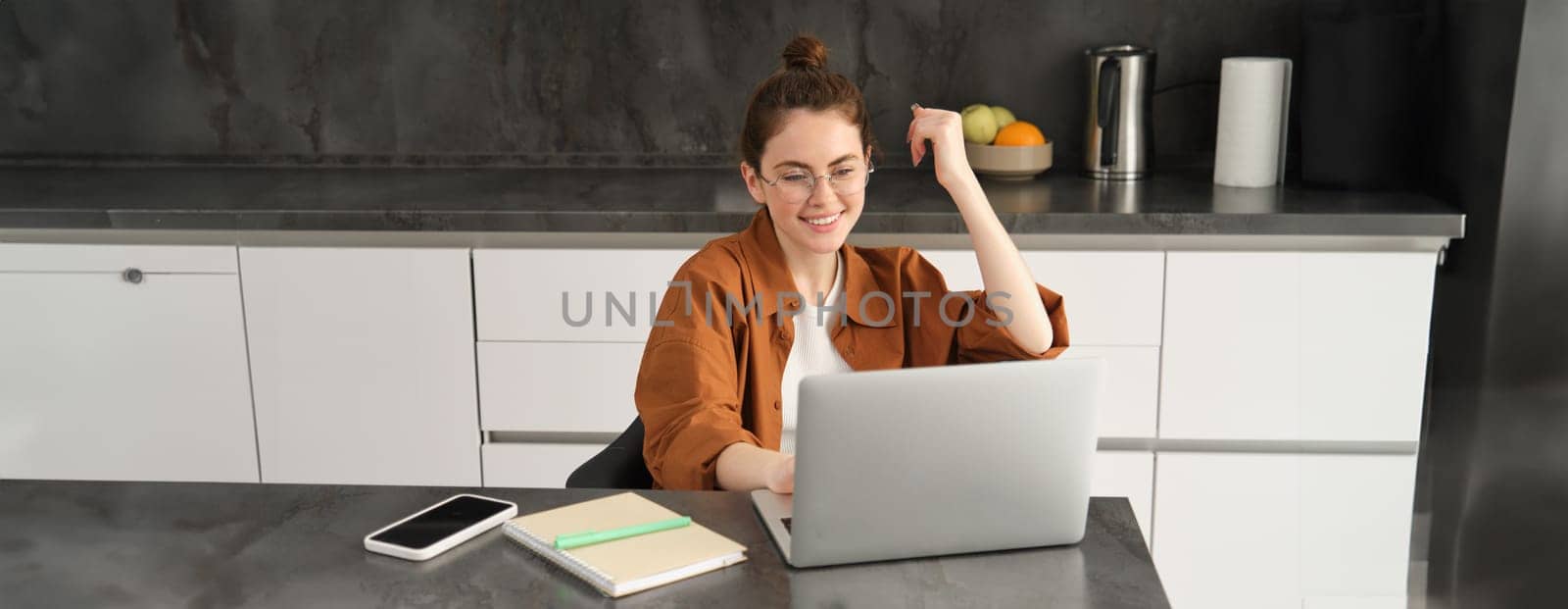 Portrait of young woman, business owner in glasses, sitting with laptop in kitchen, working from home. Student studying, doing homework on computer by Benzoix