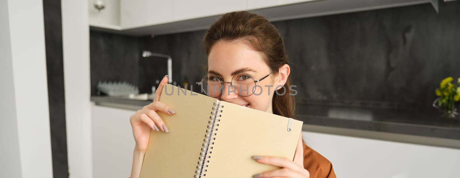 Close up portrait of smiling beautiful woman with notebook, making notes, writing in diary, studying at home, preparing homework at in kitchen.