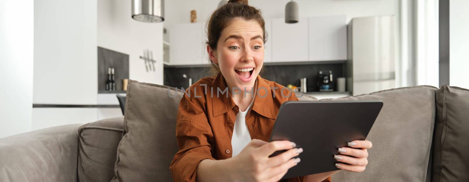 Portrait of excited, smiling young woman playing games on her digital tablet, tilting gadget and looking amused at screen, sitting on couch at home by Benzoix