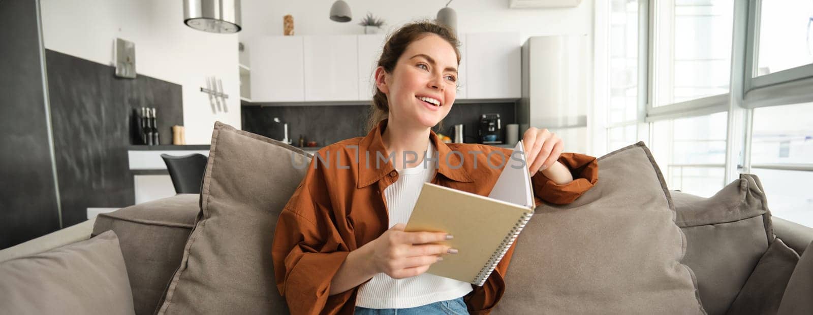 Portrait of young smiling beautiful woman, sitting on sofa in her living room, holding study material, doing homework, reading notes in notebook.