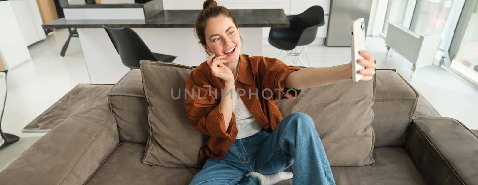 Portrait of beautiful happy girl takes selfies on sofa. Woman posing for photos on her couch, taking pictures for social media, extends hand with smartphone.