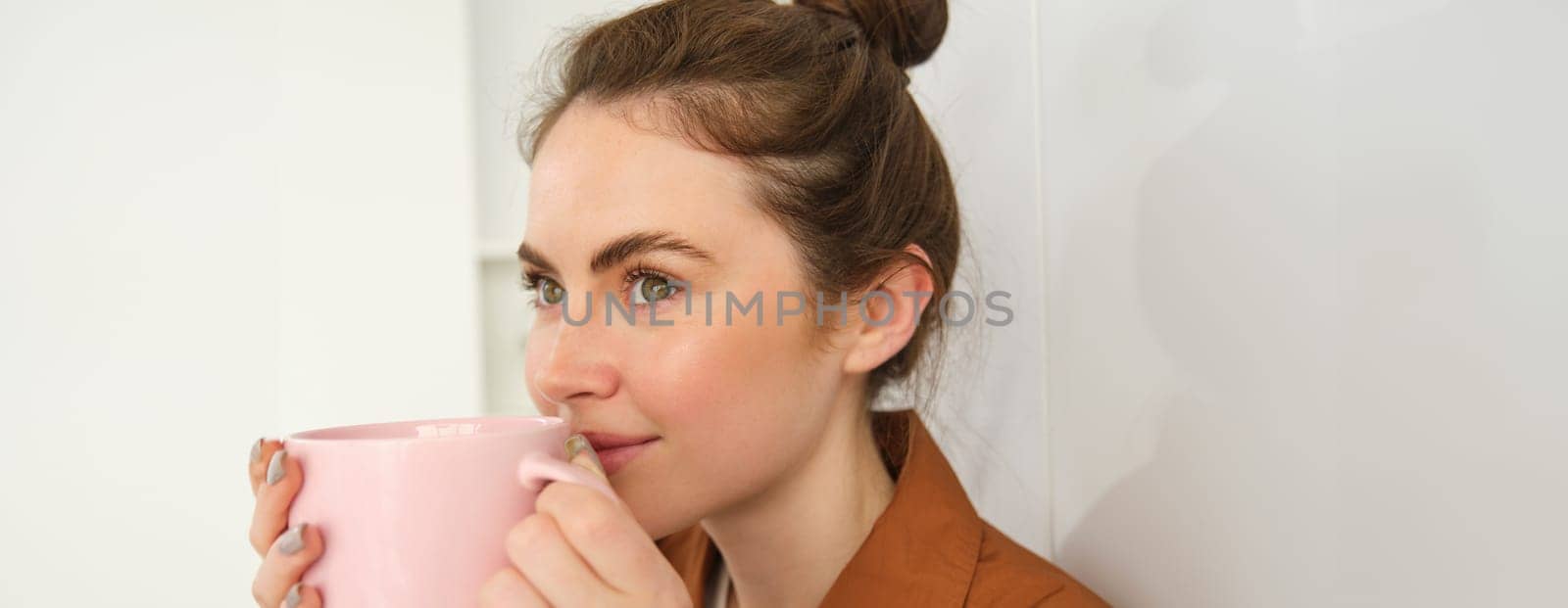 Cosy moments. Happy young woman sits in kitchen with cup of freshly brewed coffee, smiling and looking aside. Girl drinks tea at home.
