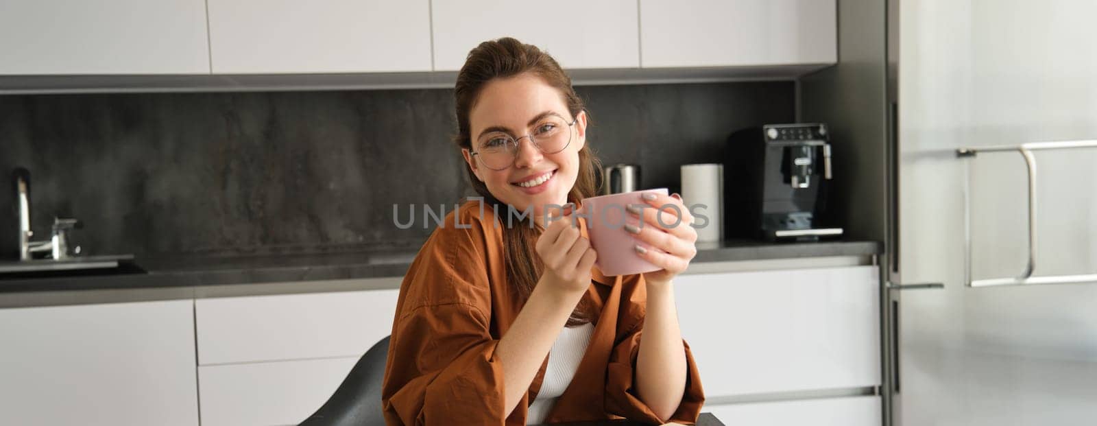 Carefree young woman in kitchen, sitting with cup of coffee and smiling at camera, drinking tea, enjoying her break time.