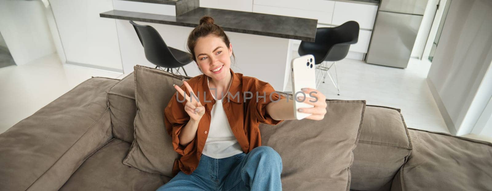 Portrait of beautiful smiling woman, takes selfie at home, posing on sofa, holds smartphone with extended hand.