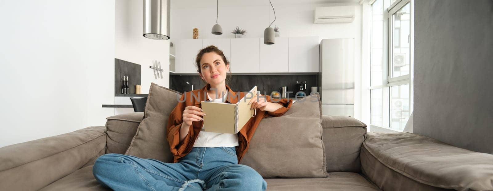 Portrait of young smiling beautiful woman, sitting on sofa in her living room, holding study material, doing homework, reading notes in notebook by Benzoix