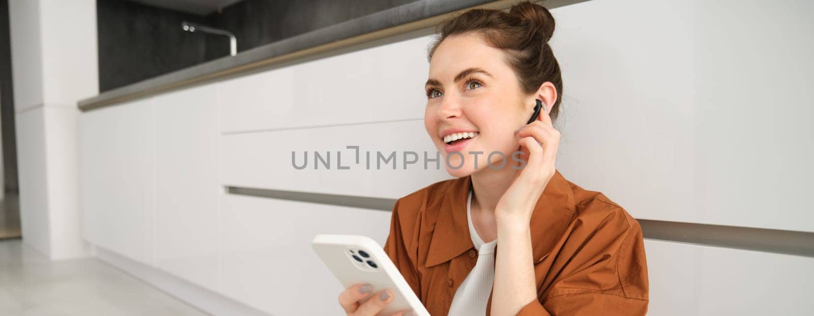 Portrait of happy young beautiful woman, listening music in wireless earphones, using smartphone, watching videos on mobile phone with headphones on by Benzoix