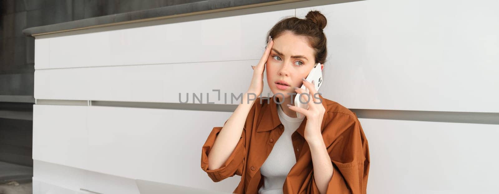 Portrait of woman calling her GP from home, holding smartphone, has headache or high fever by Benzoix