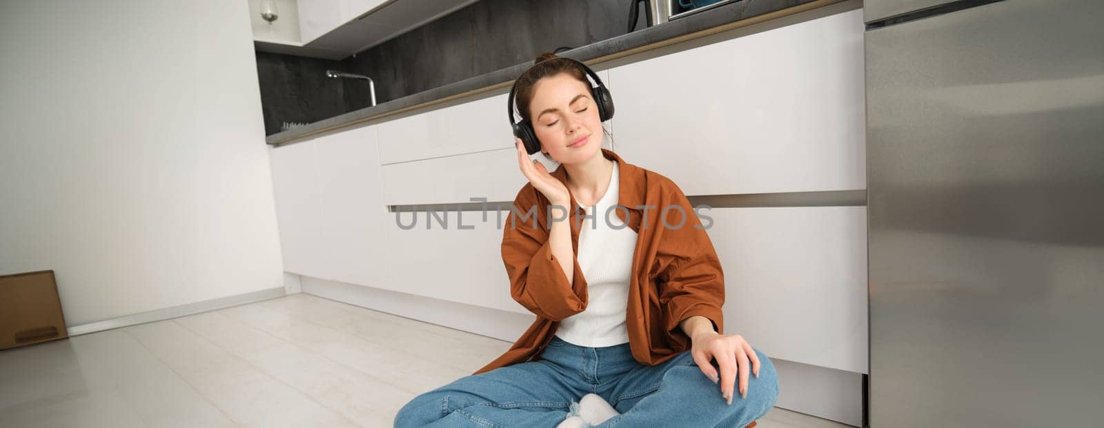 Technology concept. Young smiling brunette woman in wireless headphones, listens to music, sits on kitchen floor.