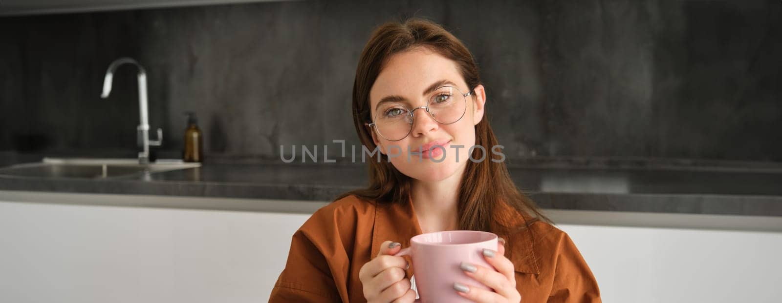 Portrait of lovely, tender young woman in glasses, sits at home with cup of tea. Girl in kitchen, enjoys drinking aromatic coffee.