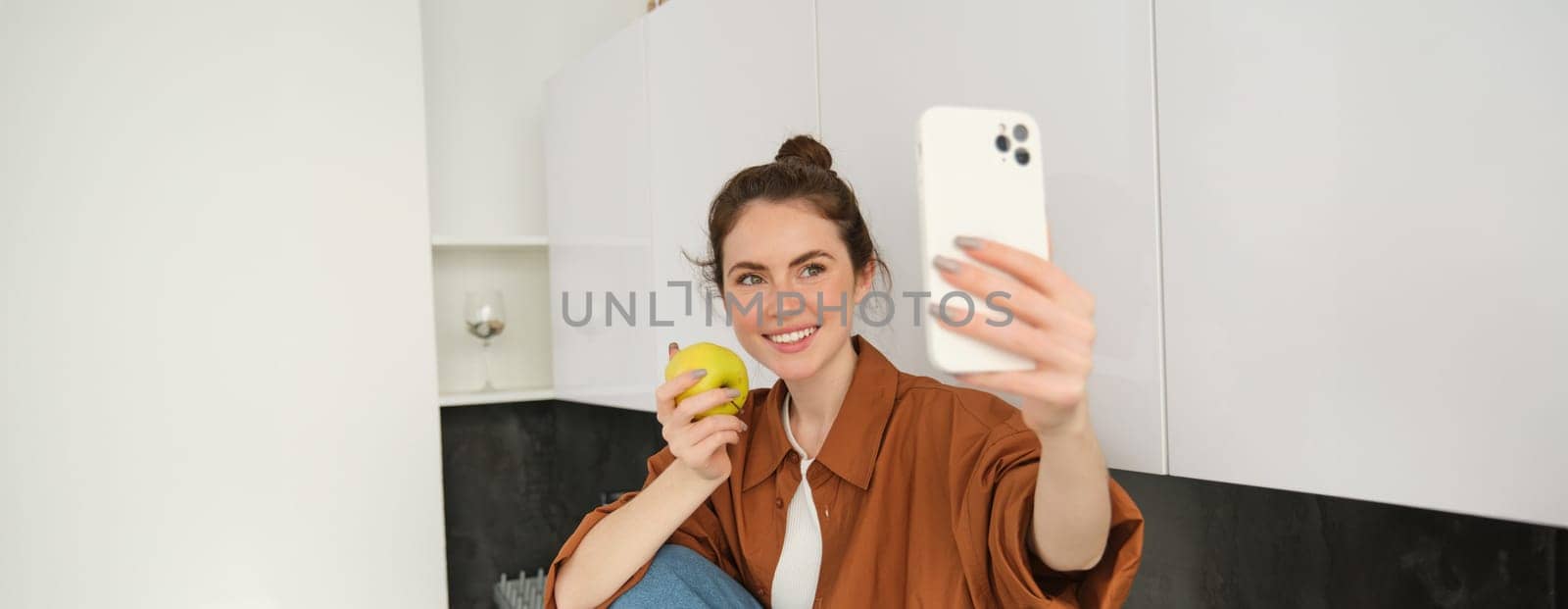 Portrait of happy, smiling young woman records herself, takes selfie while eating an apple in the kitchen, using smartphone app, makes photos with mobile phone by Benzoix