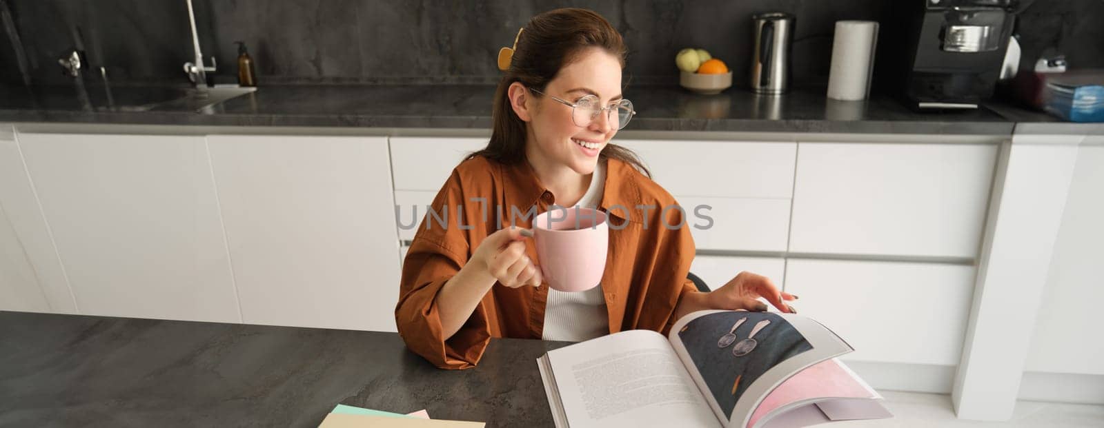 Portrait of young woman enjoying her weekend, reading a book and drinking tea at home, wearing glasses and casual clothes.