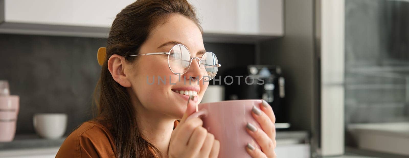Close up portrait of smiling, happy young woman in glasses, looking away, resting at home, drinking tea or coffee at home.
