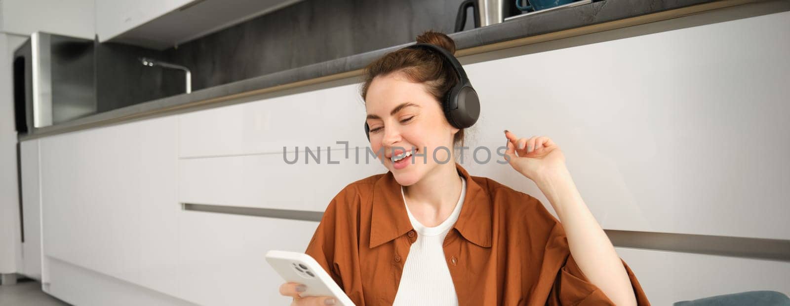 Happy woman enjoys favourite songs in her playlist, sits on floor, dances, wears headphones, listens to music.
