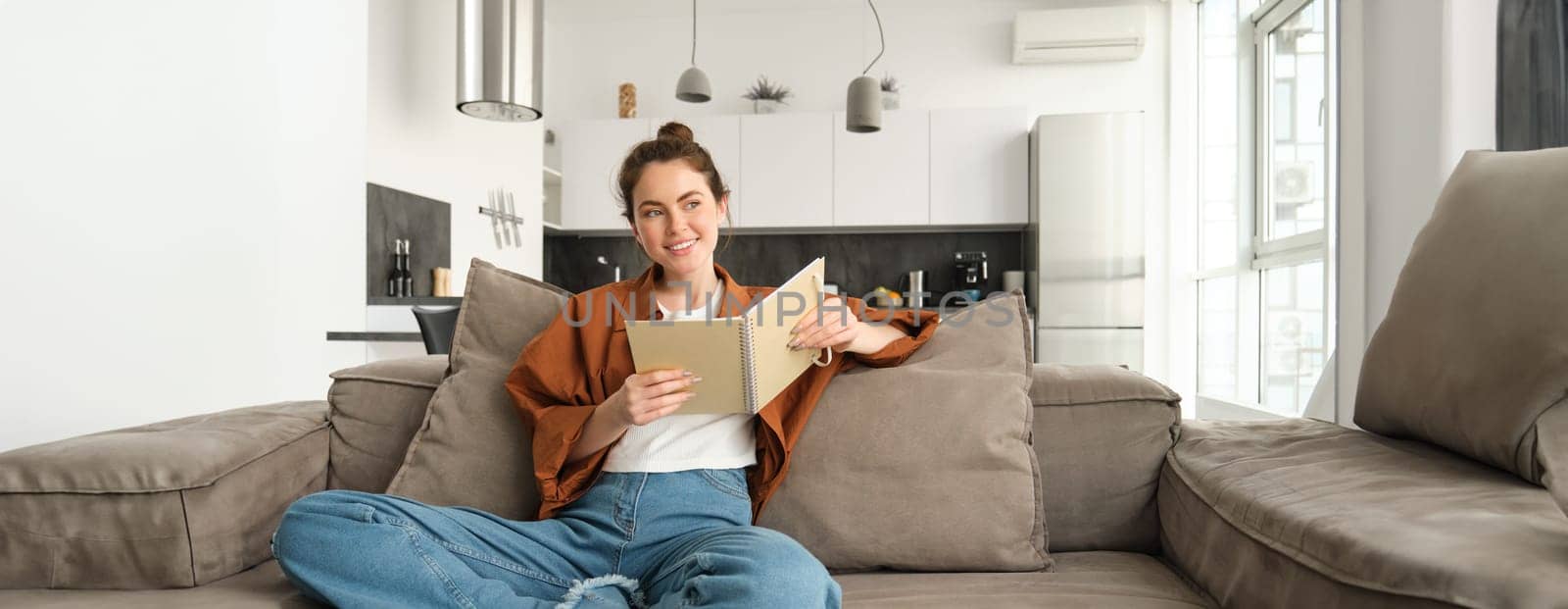 Young woman sits at home on sofa, reading her notes, holding notebook, studying for exam, student revising in her living room.