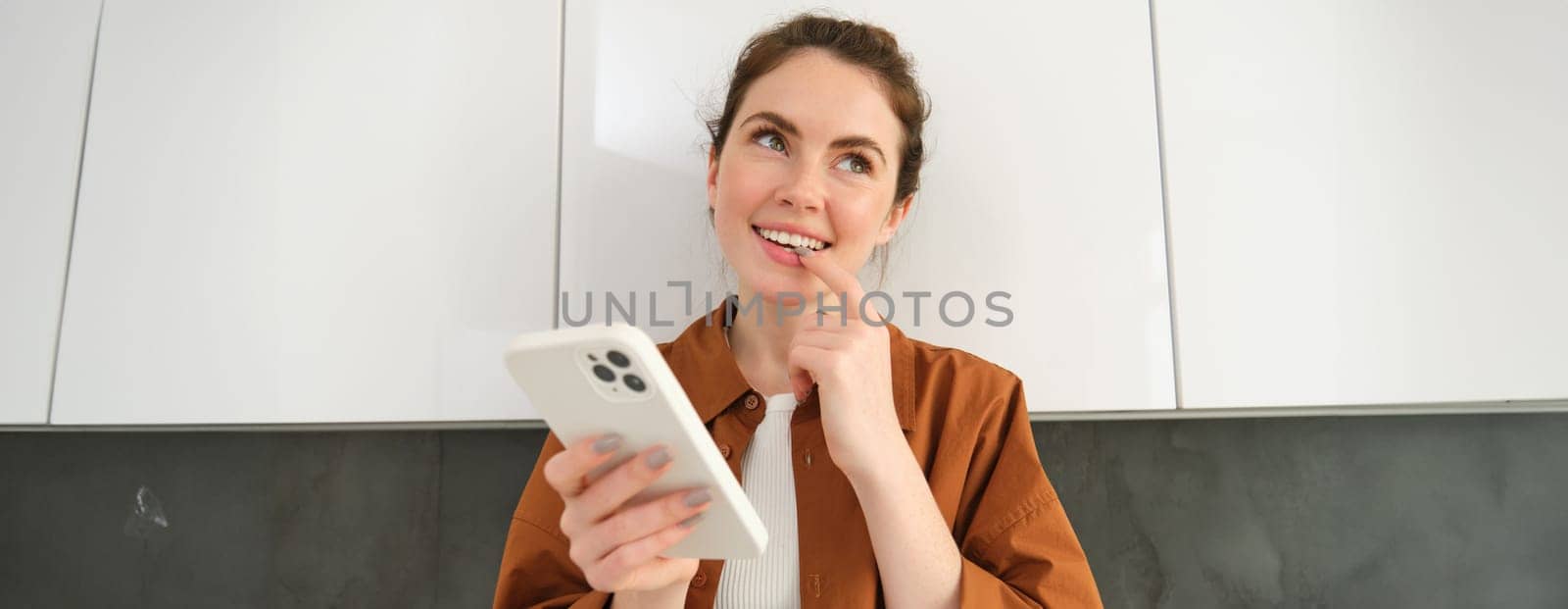 Portrait of excited young woman, smiling and looking with thinking face while using shopping app, holding smartphone.