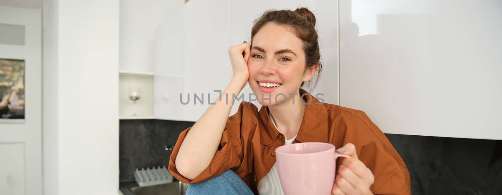 Beautiful young female model holding pink cup, sitting in kitchen and smiling at camera, woman drinking tea at home or enjoying aroma of coffee.