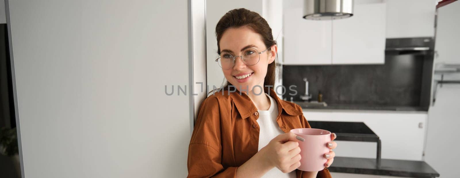 Portrait of beautiful, smiling female model, woman in glasses with cup of coffee, leans on wall at home, drinks tea.
