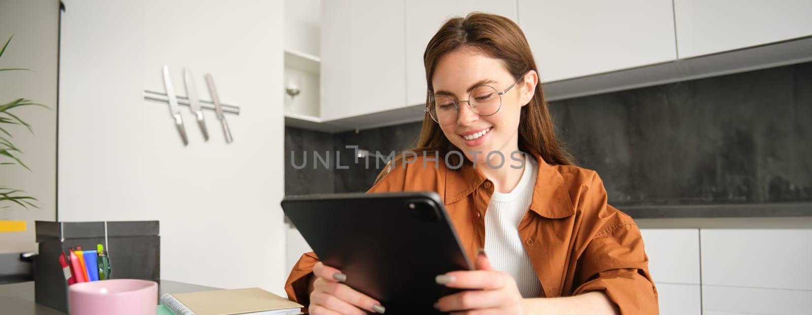 Portrait of young student, woman studying at home, working remotely, setup workplace in her kitchen, sitting on chair with digital tablet, reading in glasses by Benzoix